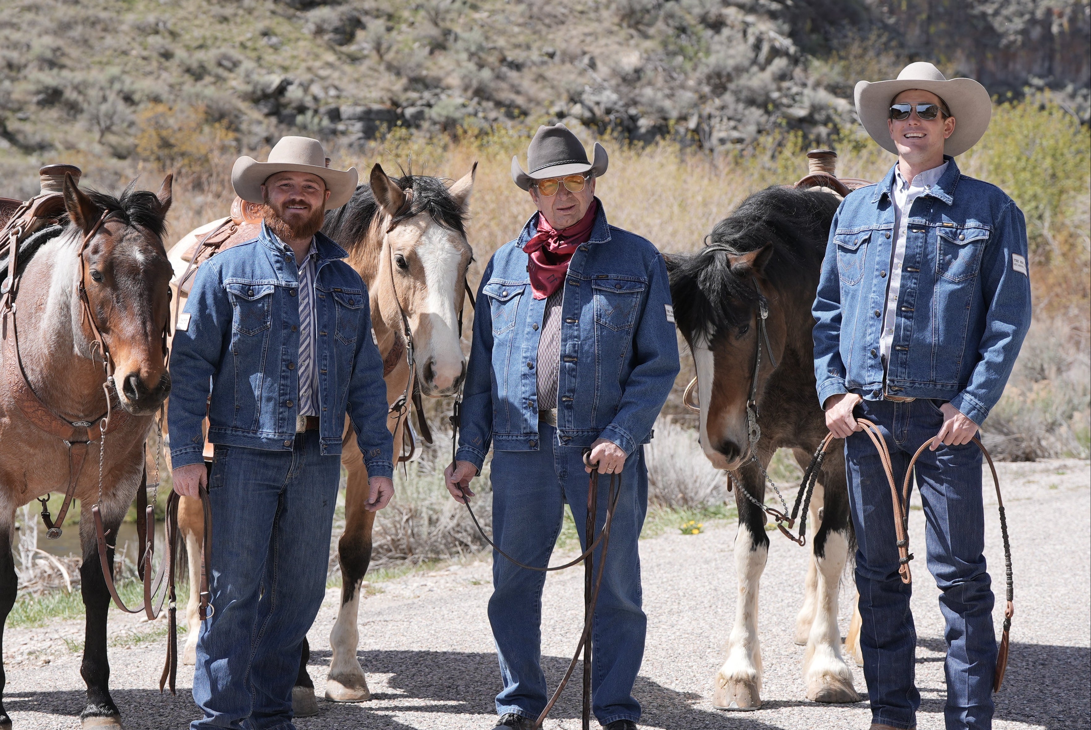 Three men in cowboy hats and jeans standing with horses in a desert landscape.