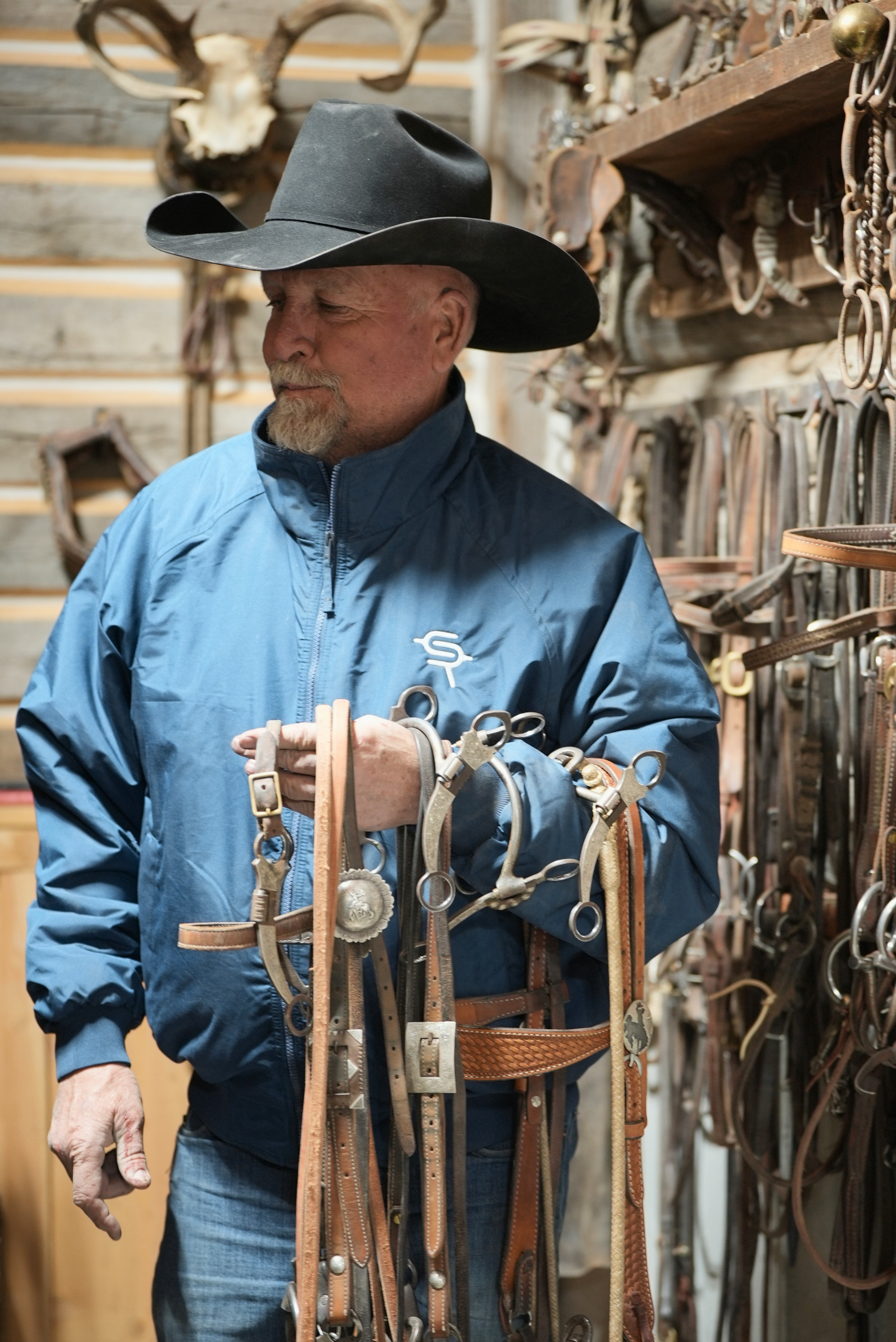 Man in a blue jacket featuring Silver Spur Ranch brand and cowboy hat holding horse reins in a tack room.