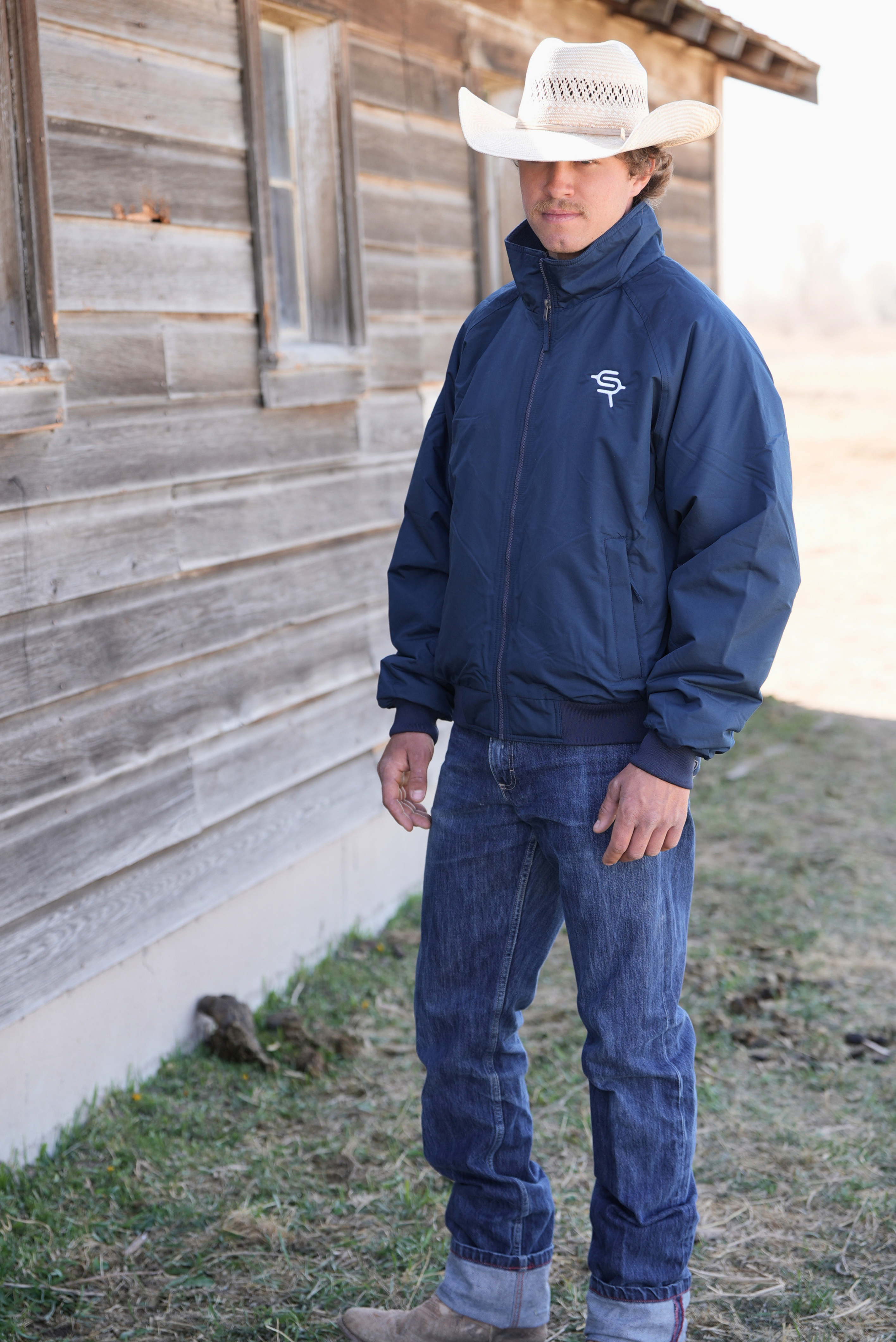 Man wearing a navy jacket with a logo and jeans standing in front of a wooden building.