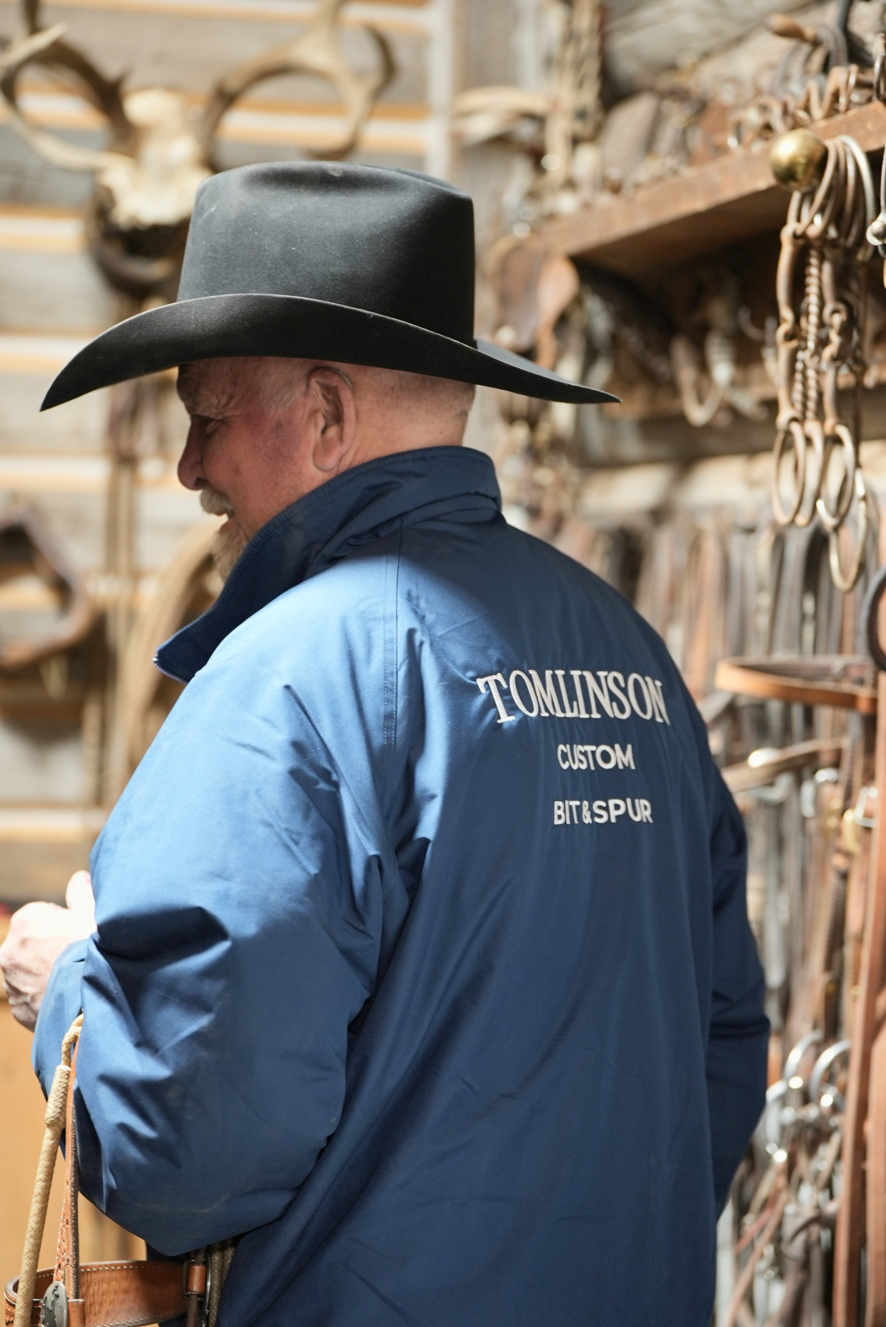 Man wearing a blue jacket with 'Tomlinson Custom Bit & Spurs' branding, standing in front of a display of cowboy hats and accessories.