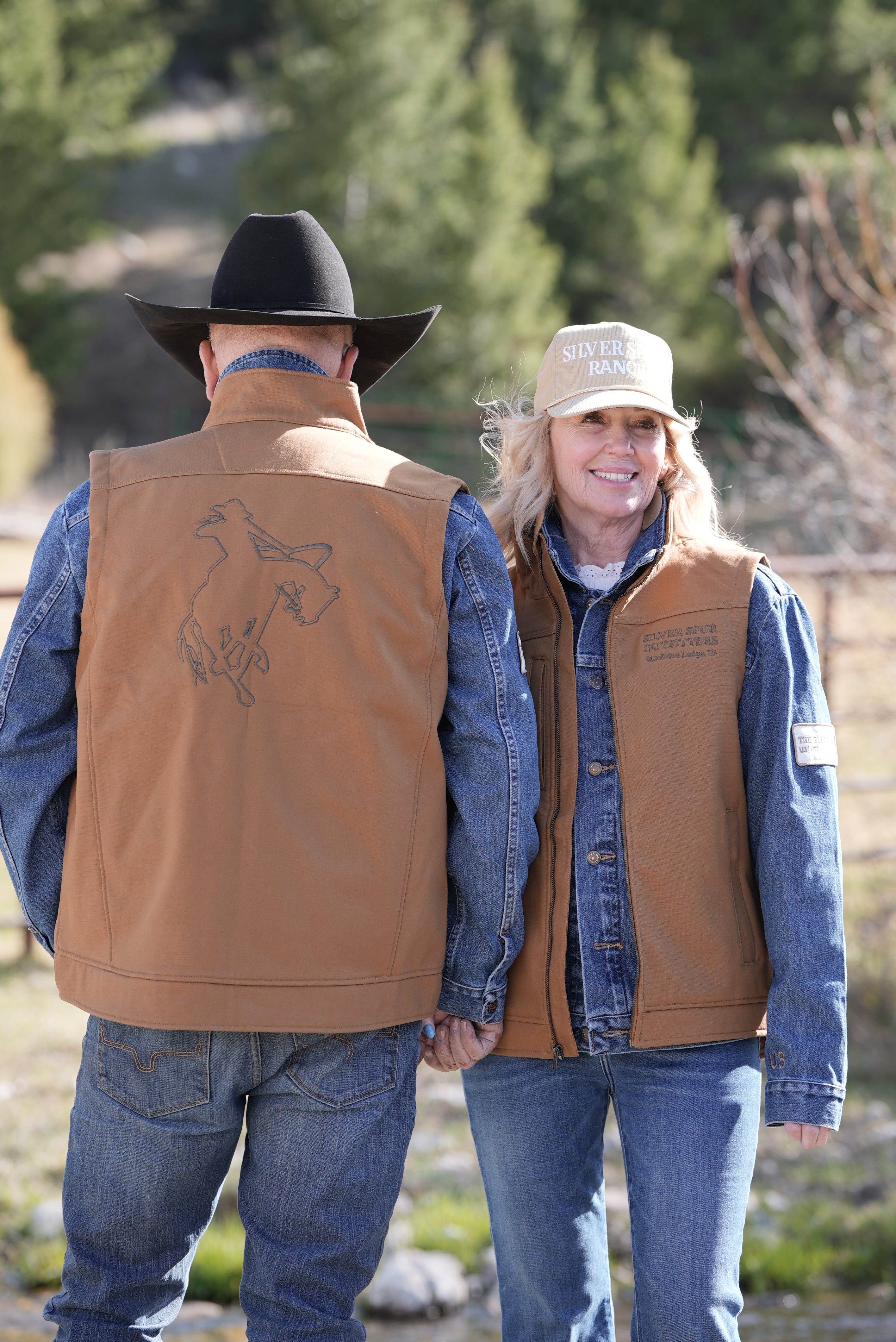 Two people wearing a brown vest featuring the Silver Spur Ranch logo, standing outdoors with trees in the background.