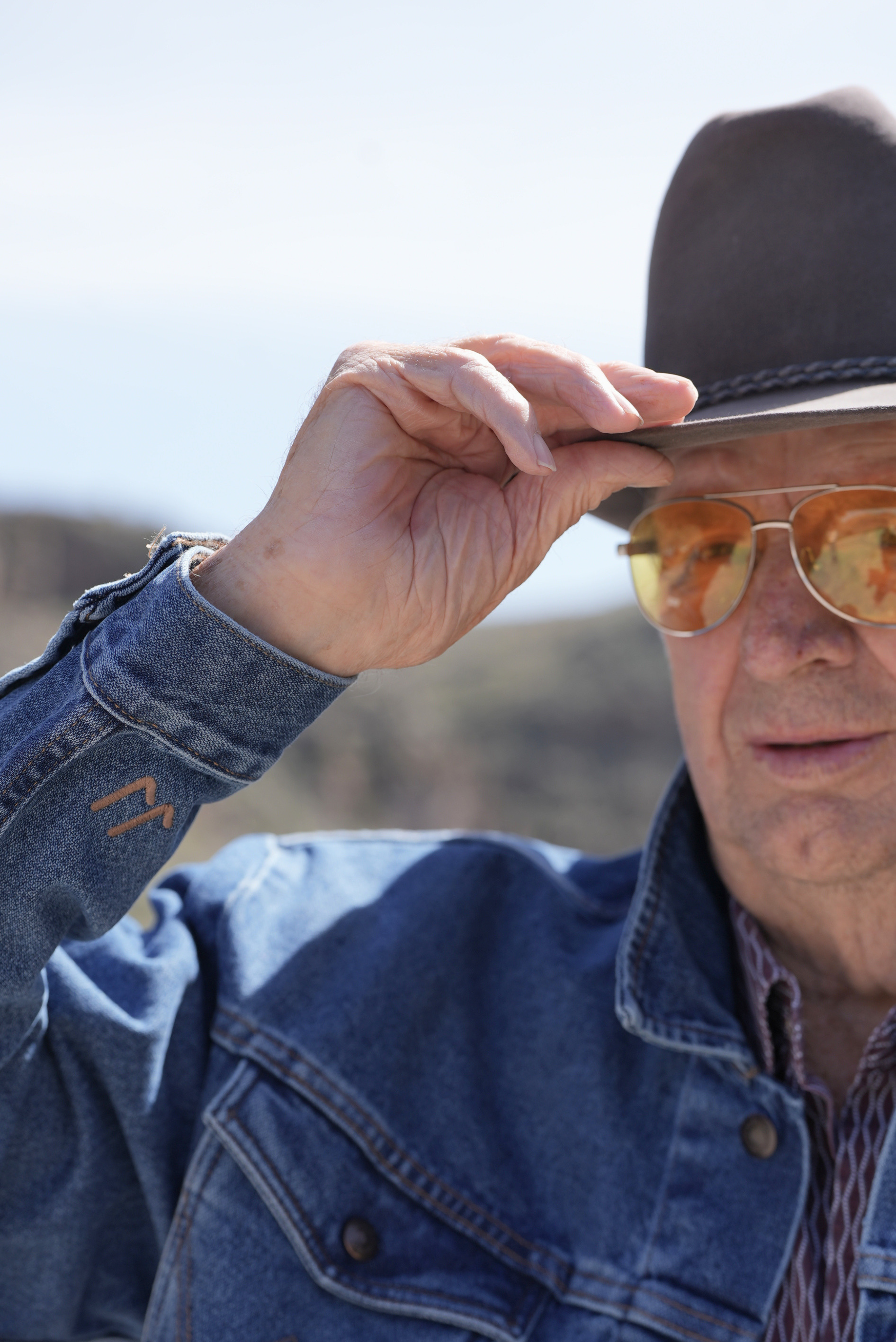 Man wearing a denim jacket, sunglasses, and a black hat outdoors.