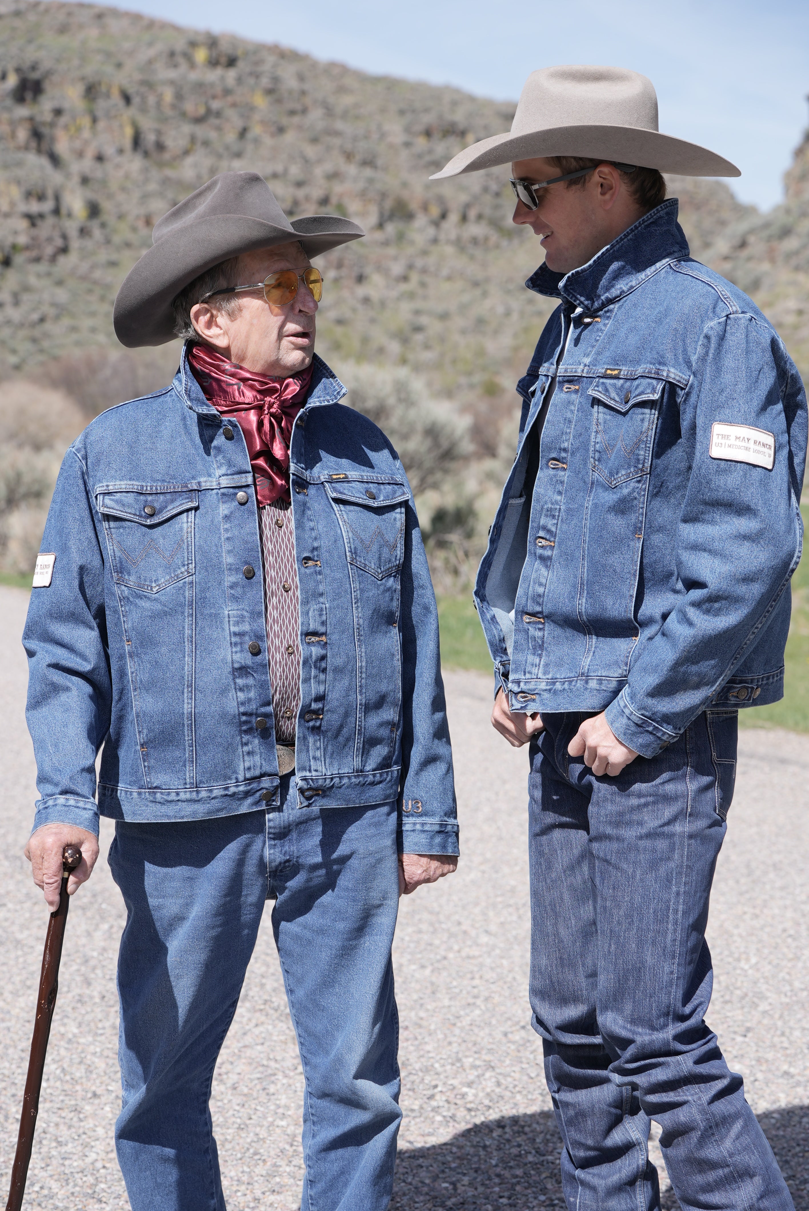 Two men in cowboy hats and denim jackets standing on a road with a mountainous landscape in the background.