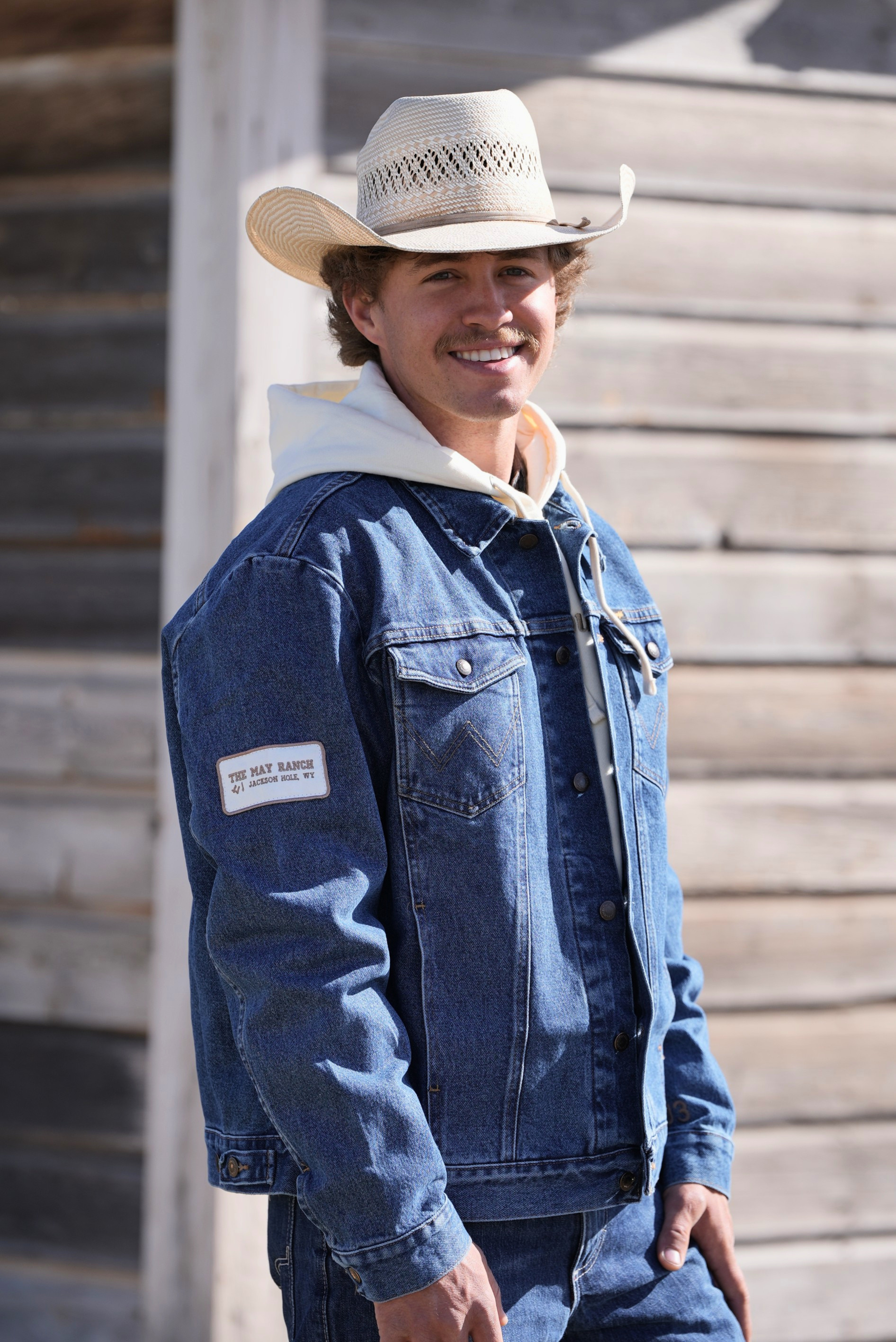 Man wearing a blue denim jacket with white fur lining and a cowboy hat, standing against a wooden background.