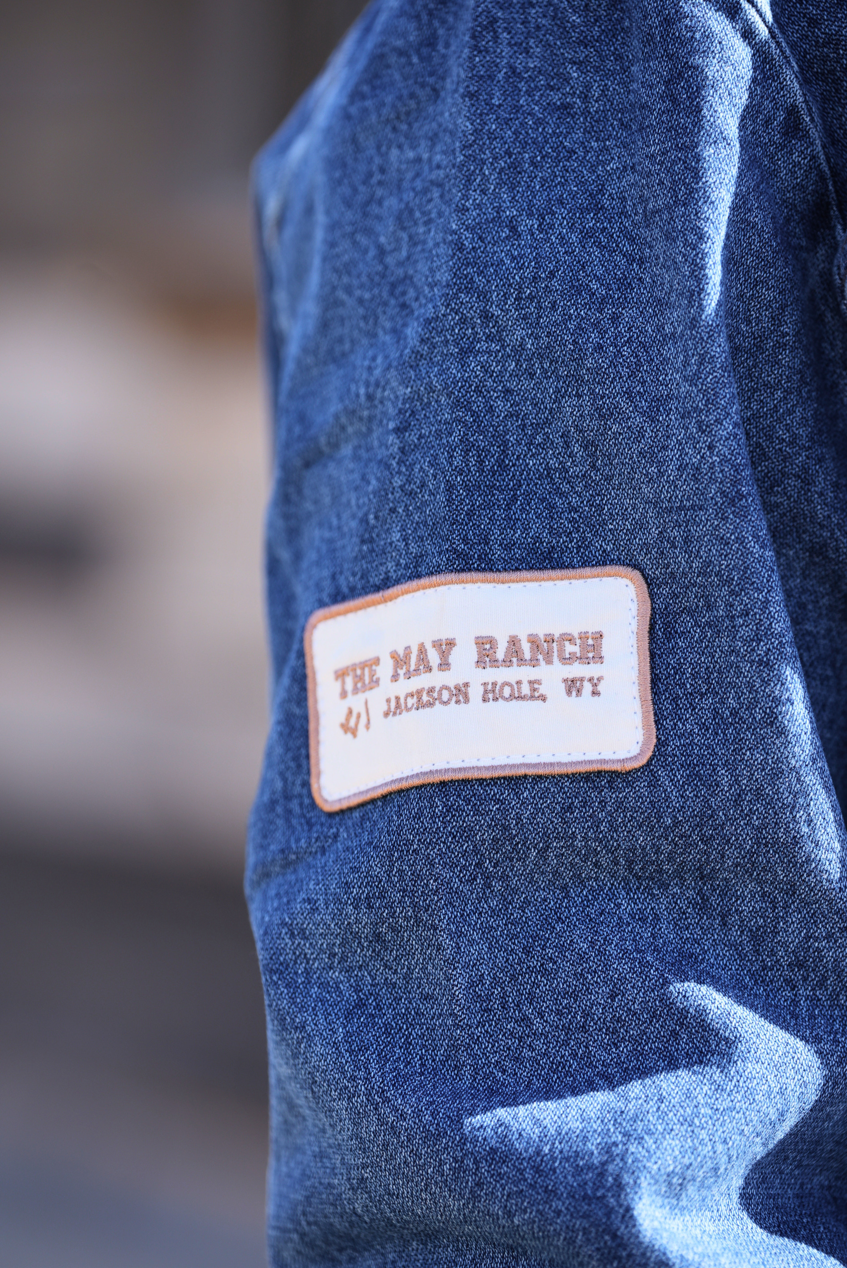 Blue jacket with a patch reading 'The May Ranch Jackson Hole, WY' on a blurred background.