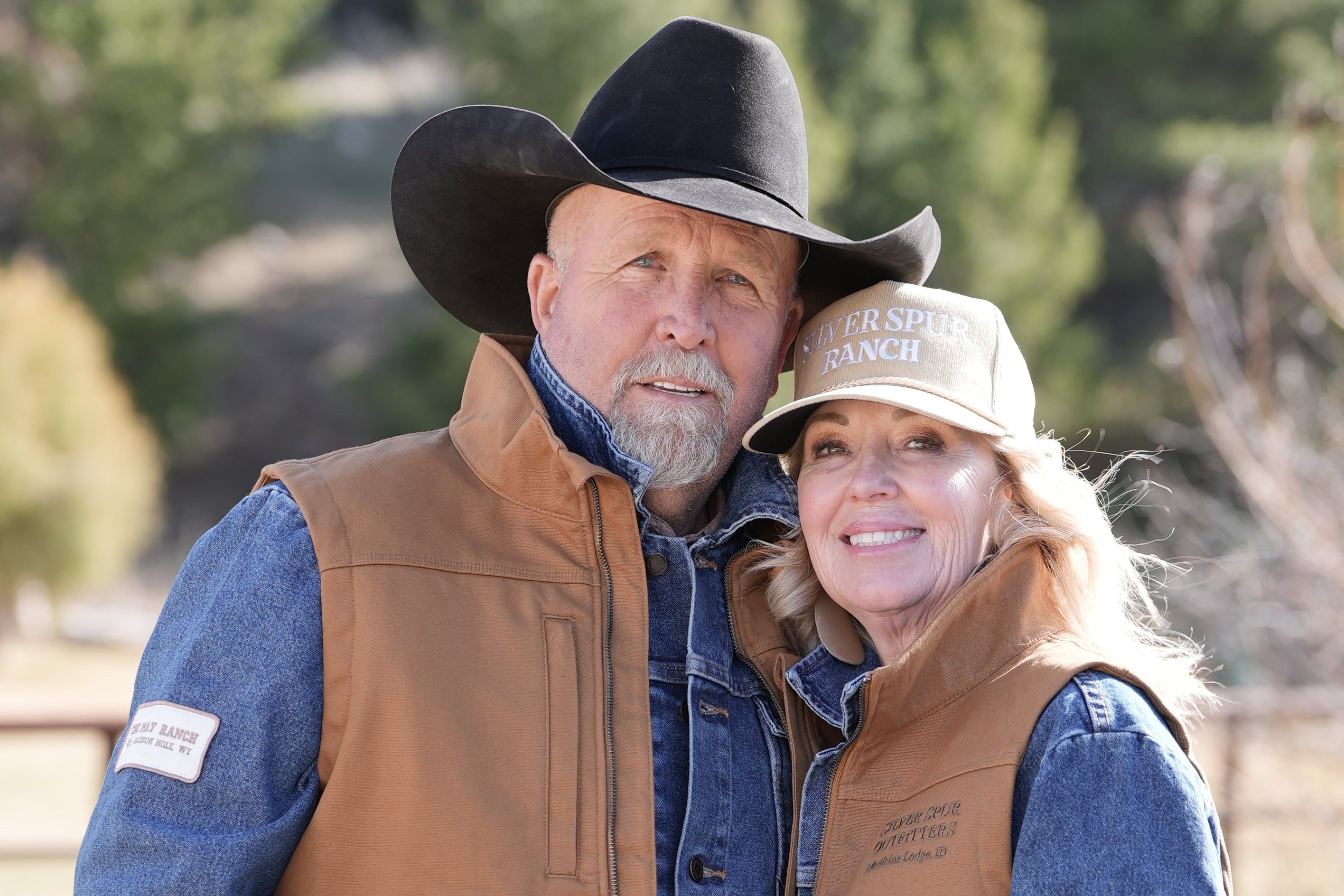Man and woman wearing cowboy hats and vests standing outdoors with trees in the background
