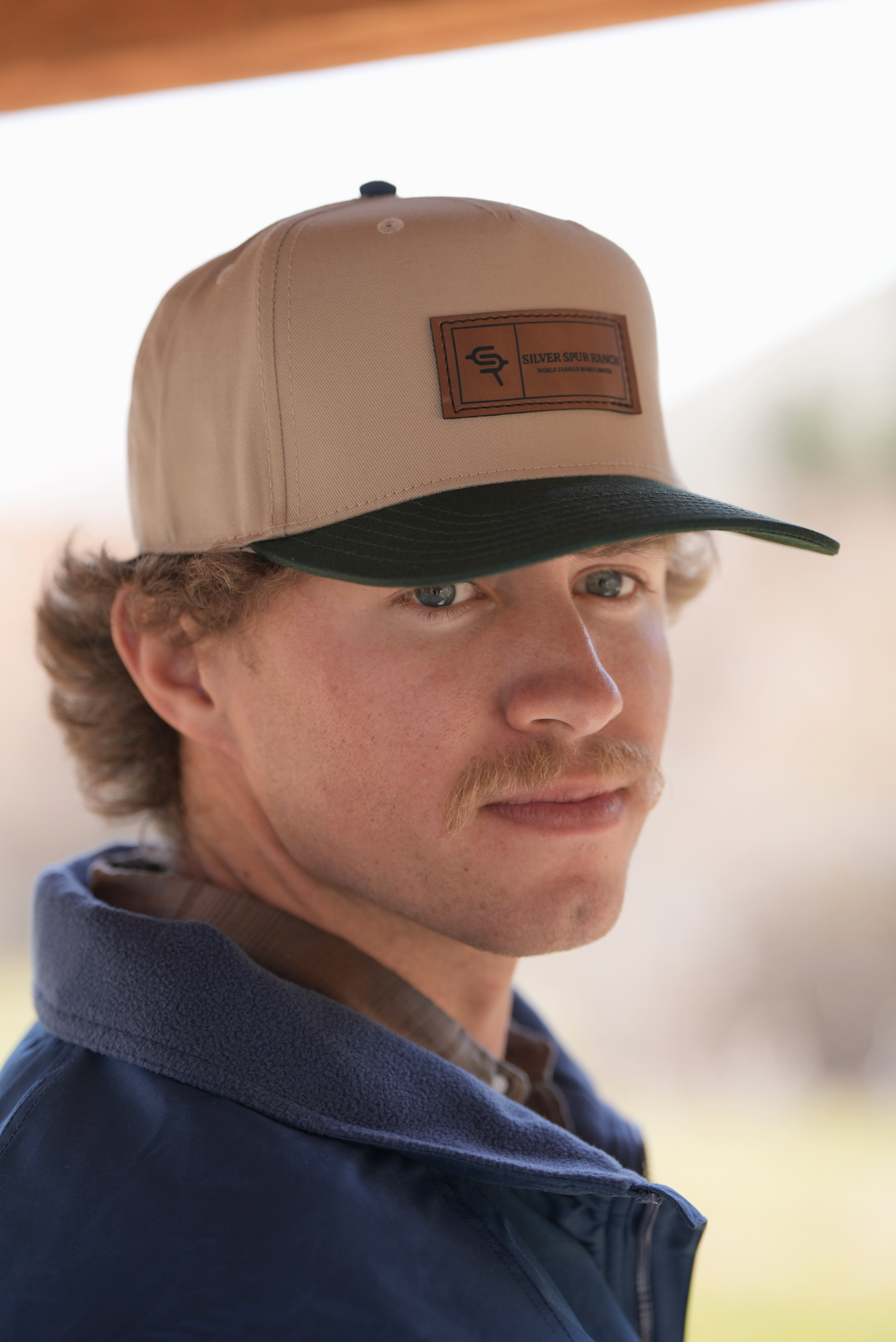 Man wearing a beige two tone cap with a leather logo, looking to the side.