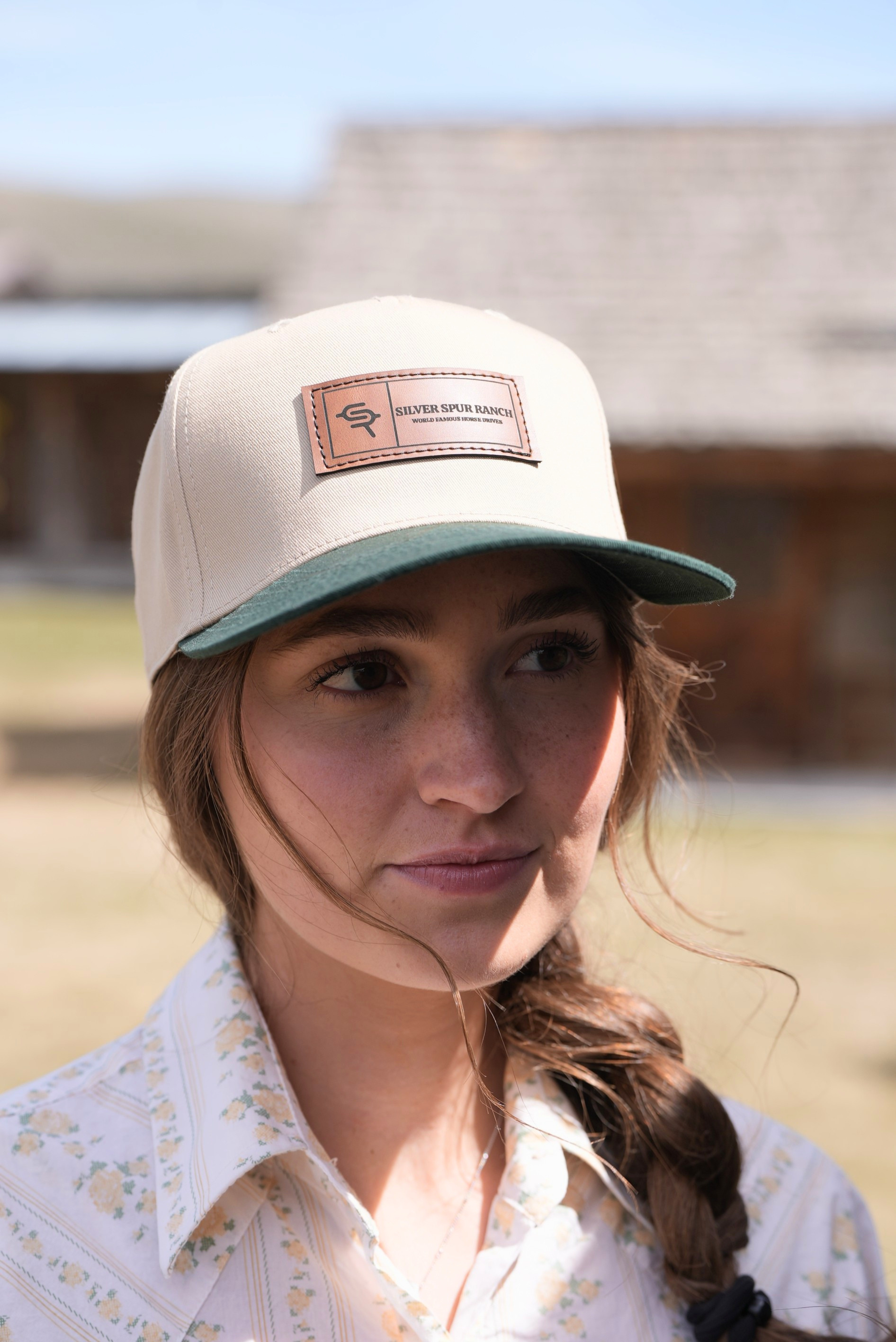 Woman wearing a beige two tone cap with a leather logo, standing outdoors.