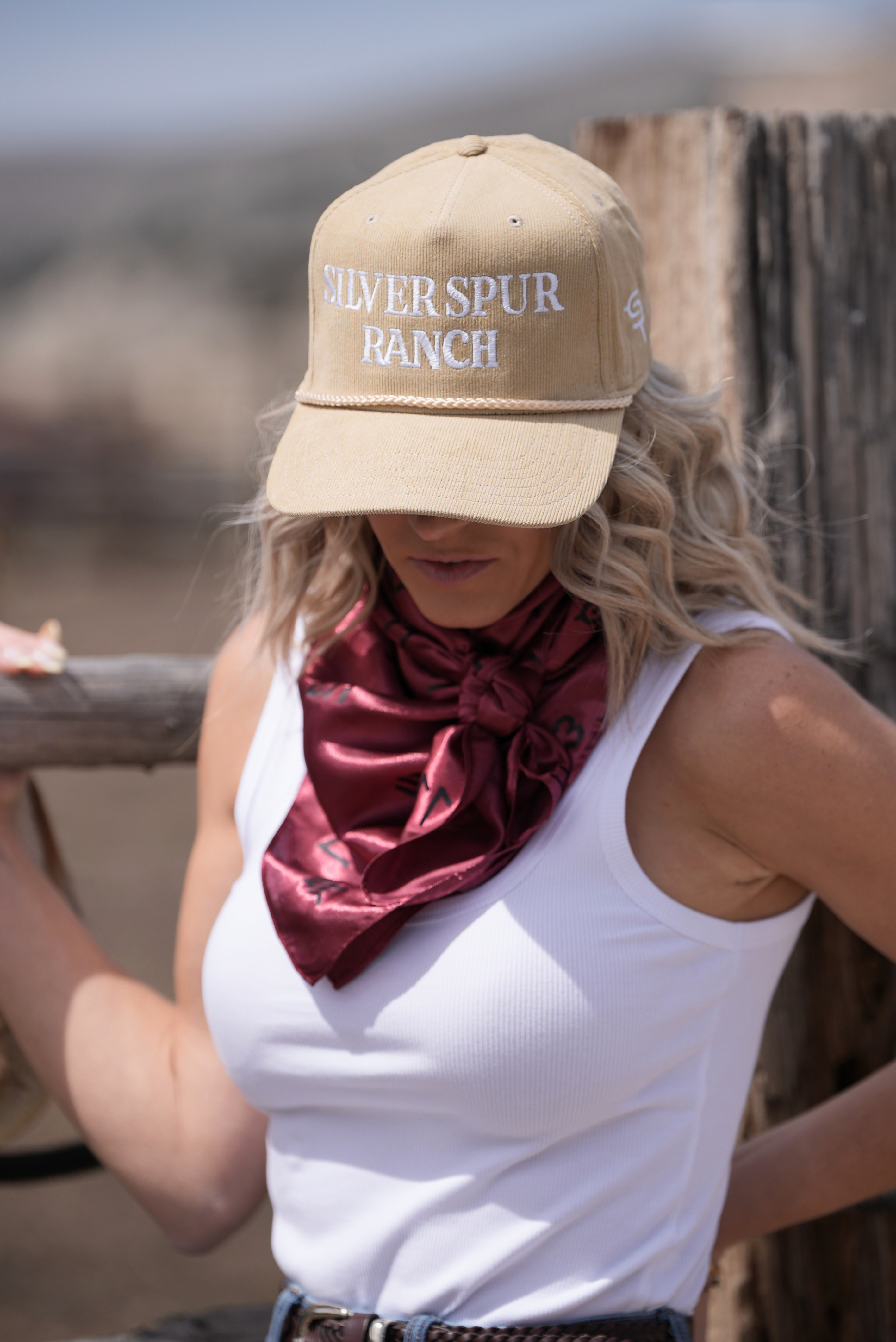 Woman wearing a red satin silk wild rag scarf standing by a fence wearing a 'Silver Spur Ranch' hat.