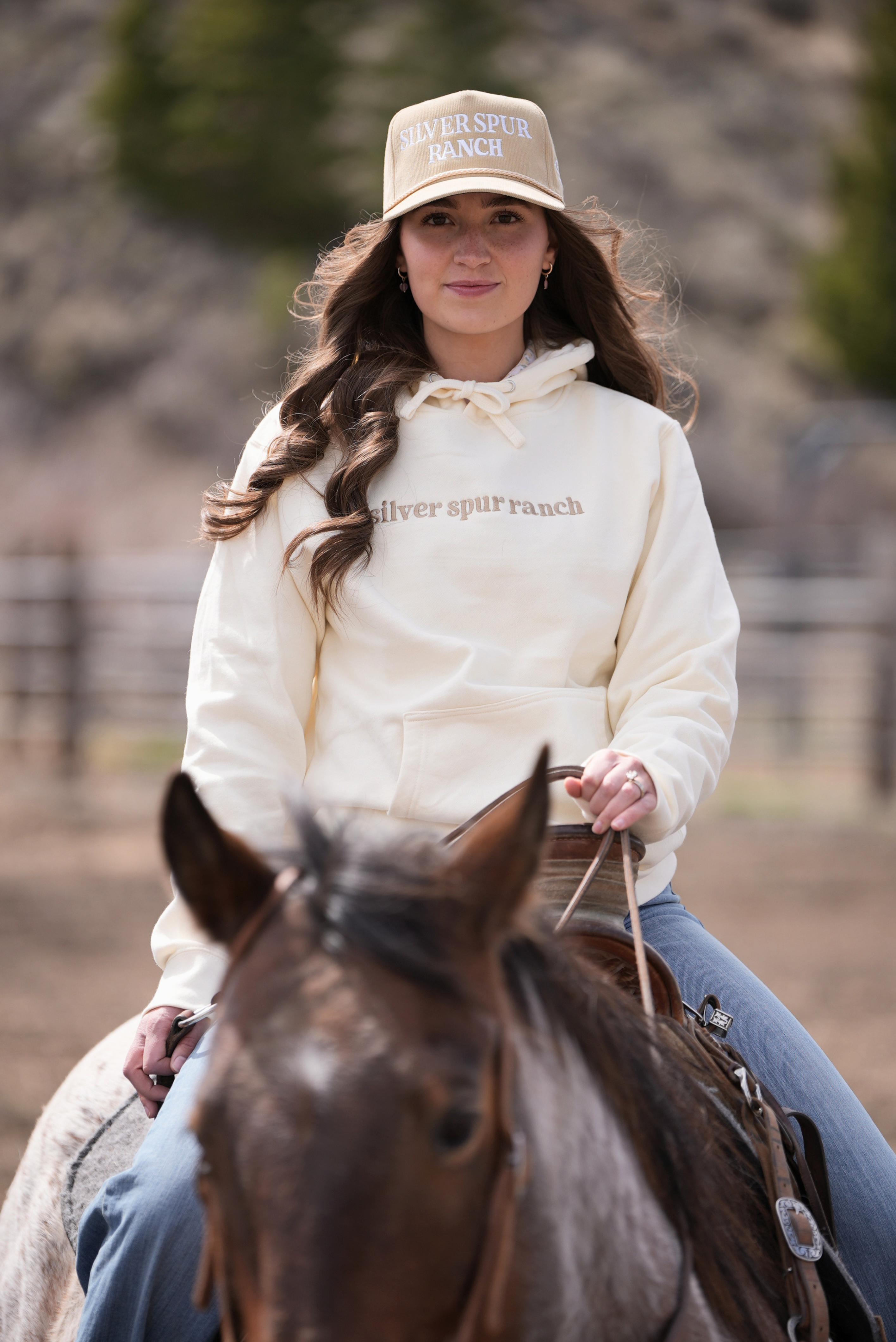 Woman riding a horse wearing a cap and hoodie with 'Silver Spurs Ranch' branding.
