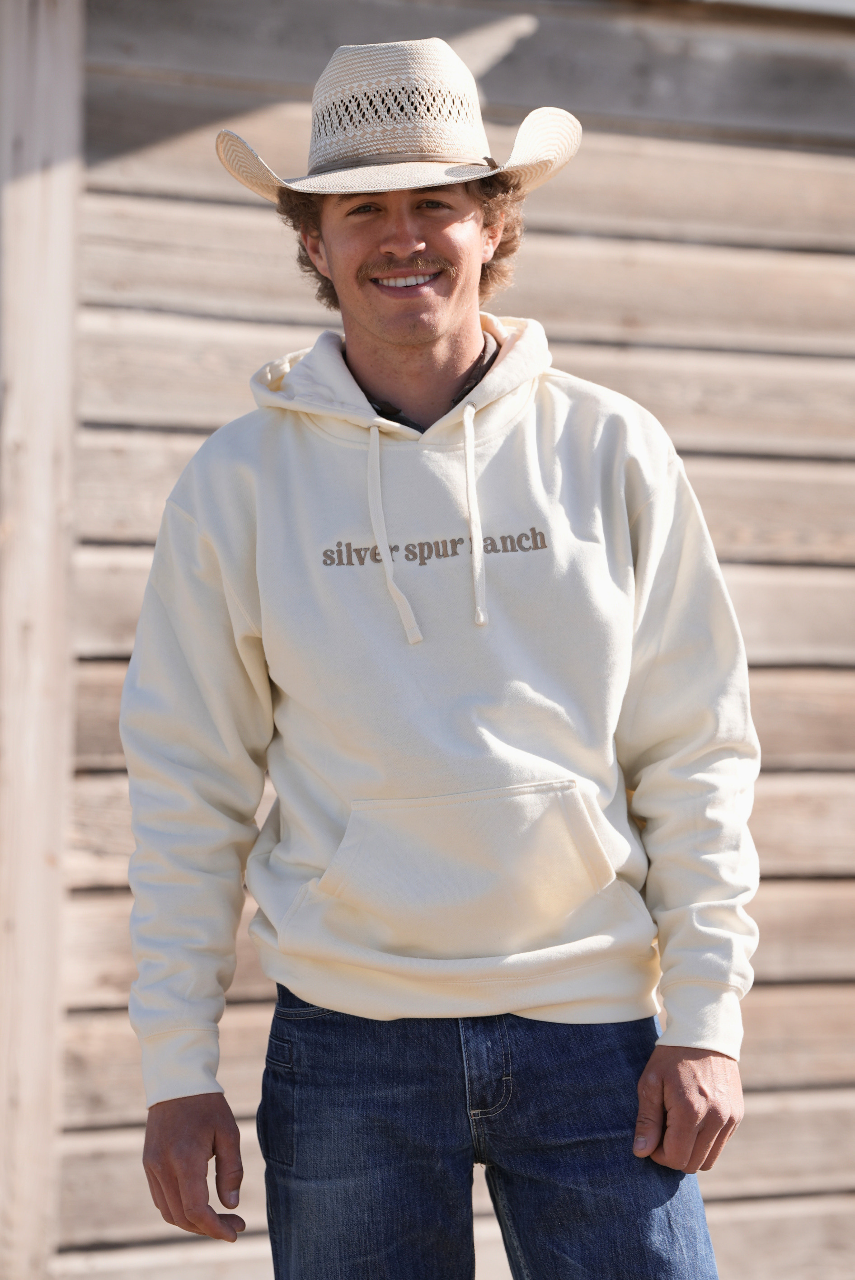 Man wearing a white hoodie with 'Silver Spurs Ranch' text, blue jeans, and a cowboy hat against a wooden background.