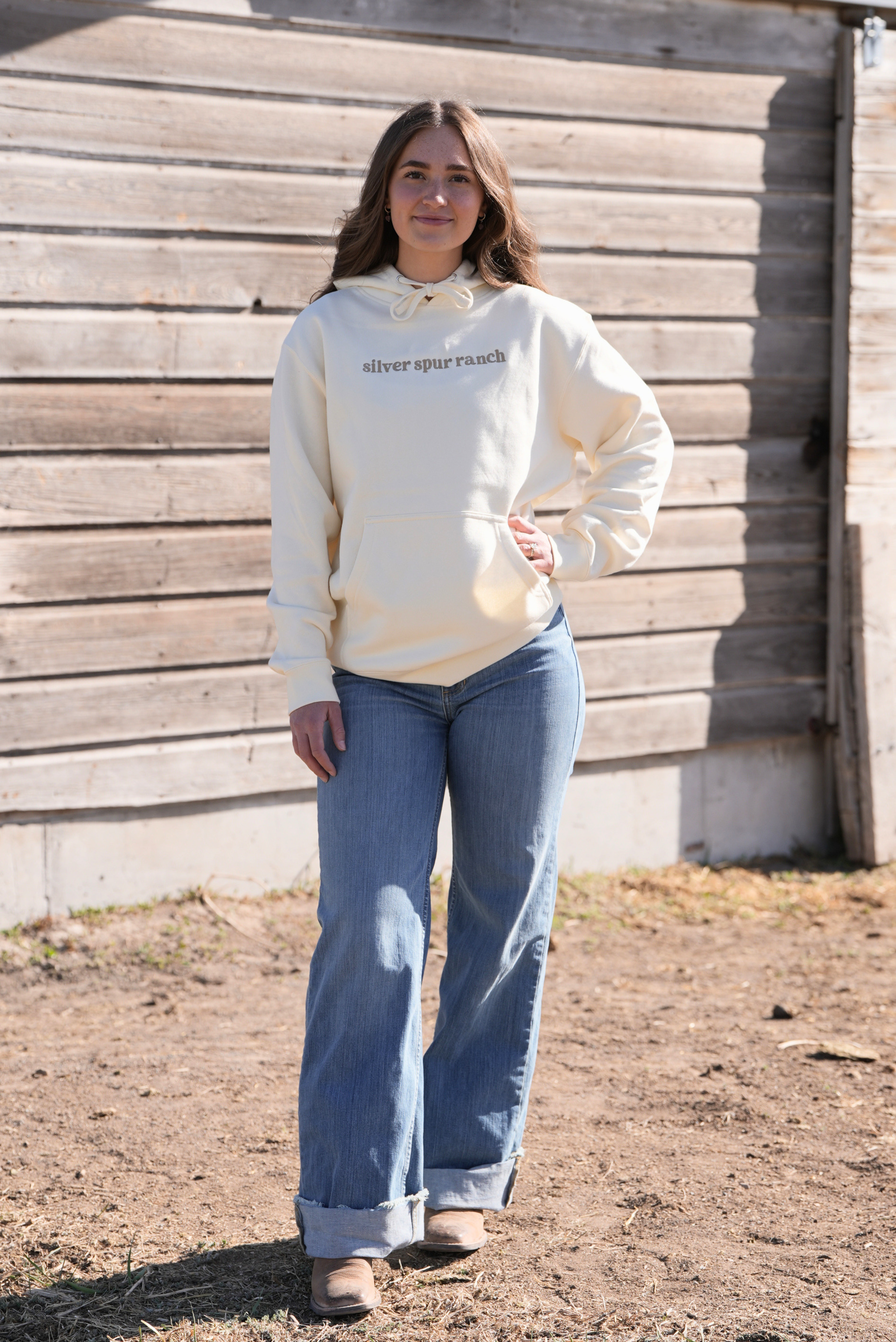 Woman wearing a cream-colored sweatshirt with text that says 'Silver Spur Ranch' and blue jeans standing in front of a wooden wall.